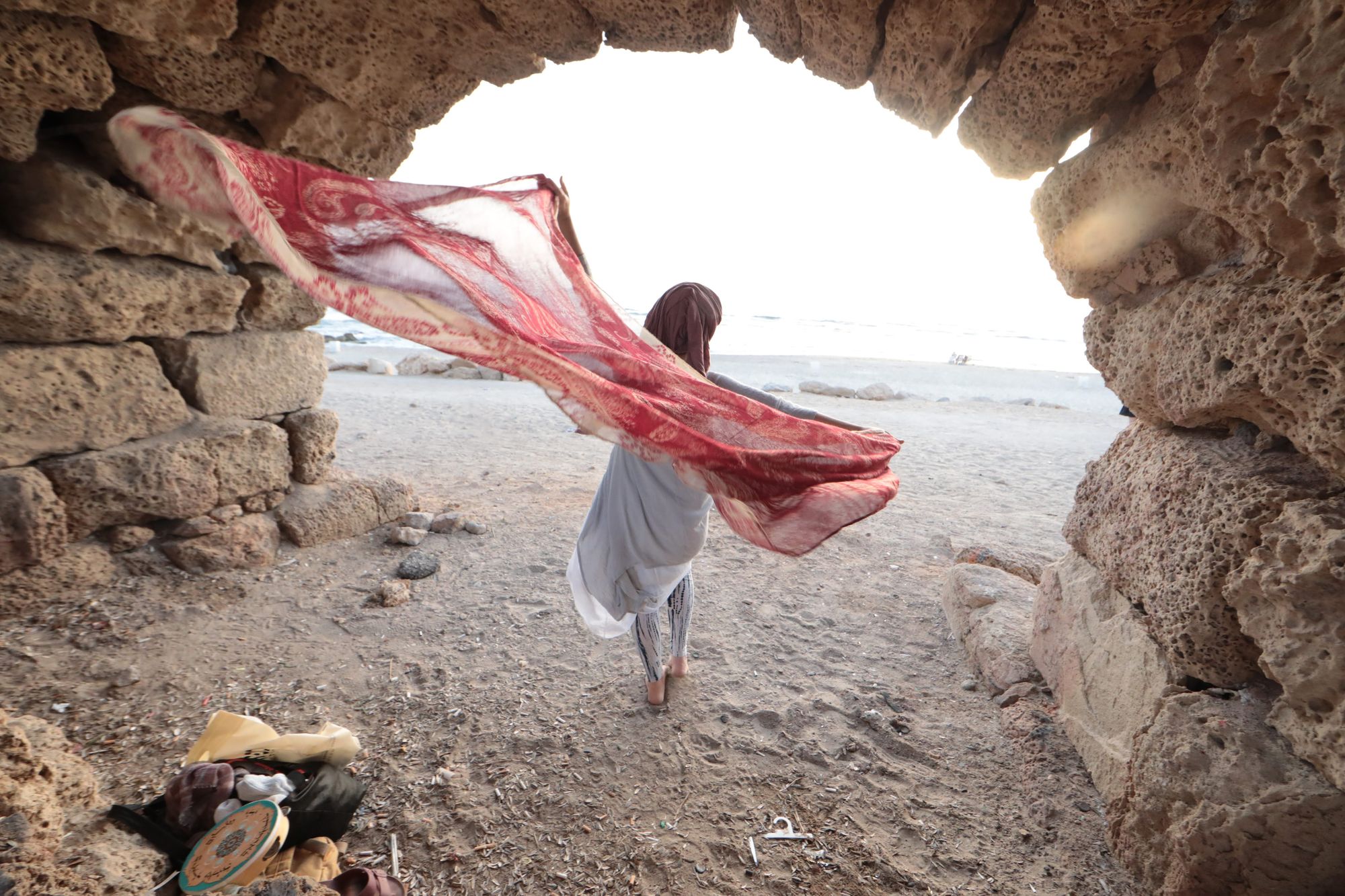 A stone arch on the shore, opening onto the sea, a woman walking out through it with a scarf spread wide in the wind